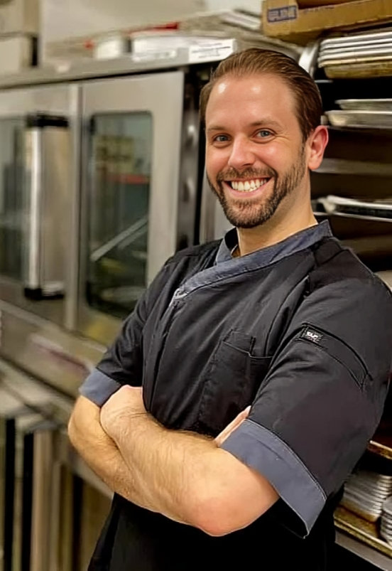 Ryan Chvala-Manges, owner and baker, smiling in a black chef coat in a kitchen